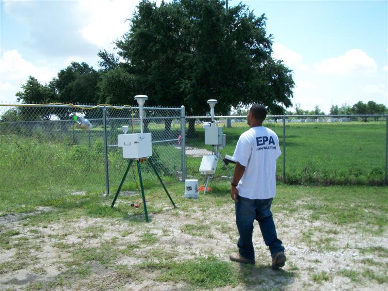 EPA air sampling instruments on tripods inside a fenced monitoring site, with a technician in an EPA contractor shirt walking past