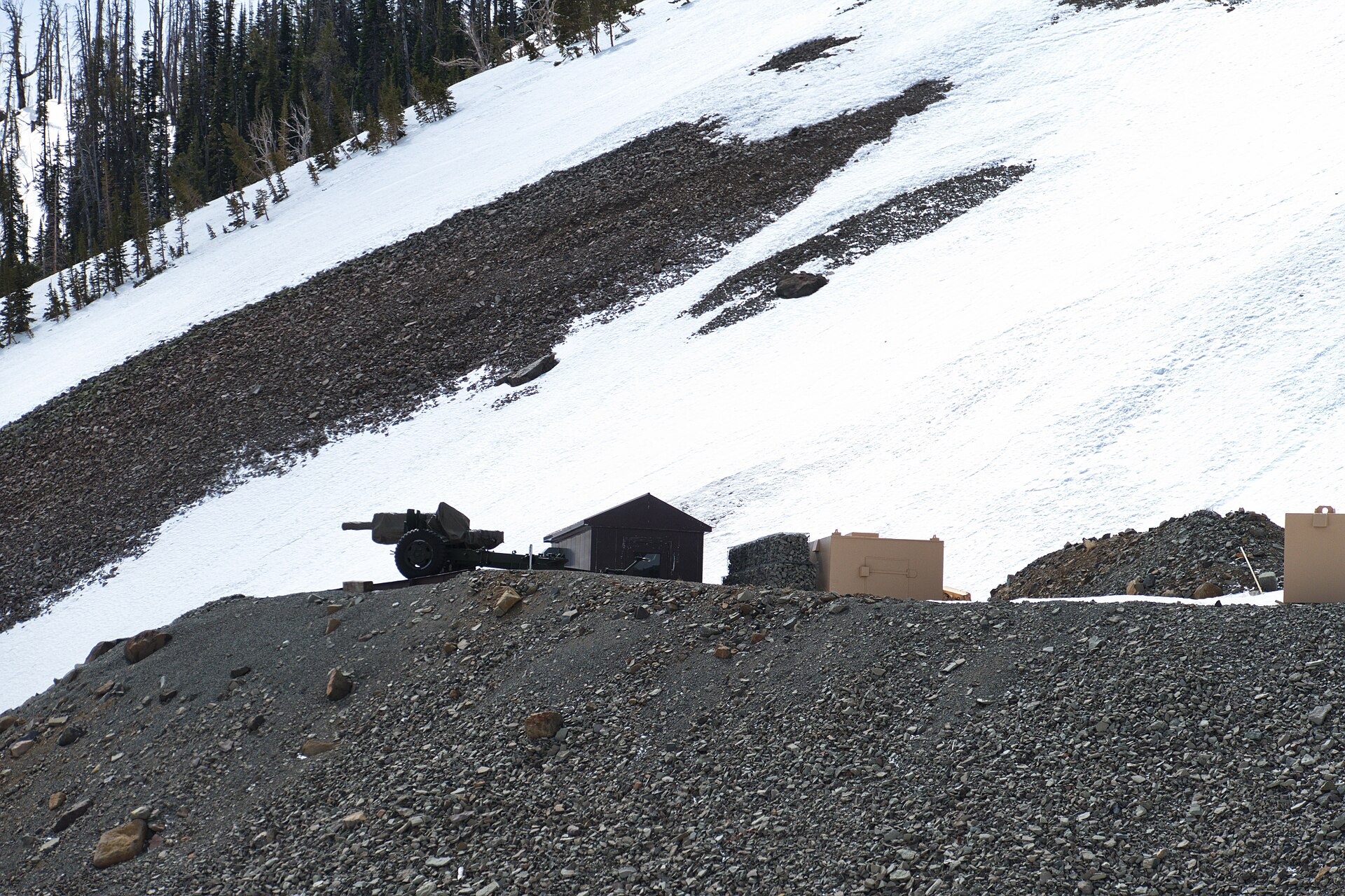 An artillery piece on a gravel berm aimed across a wide snow-covered mountainside, with a small dark hut and equipment boxes beside it