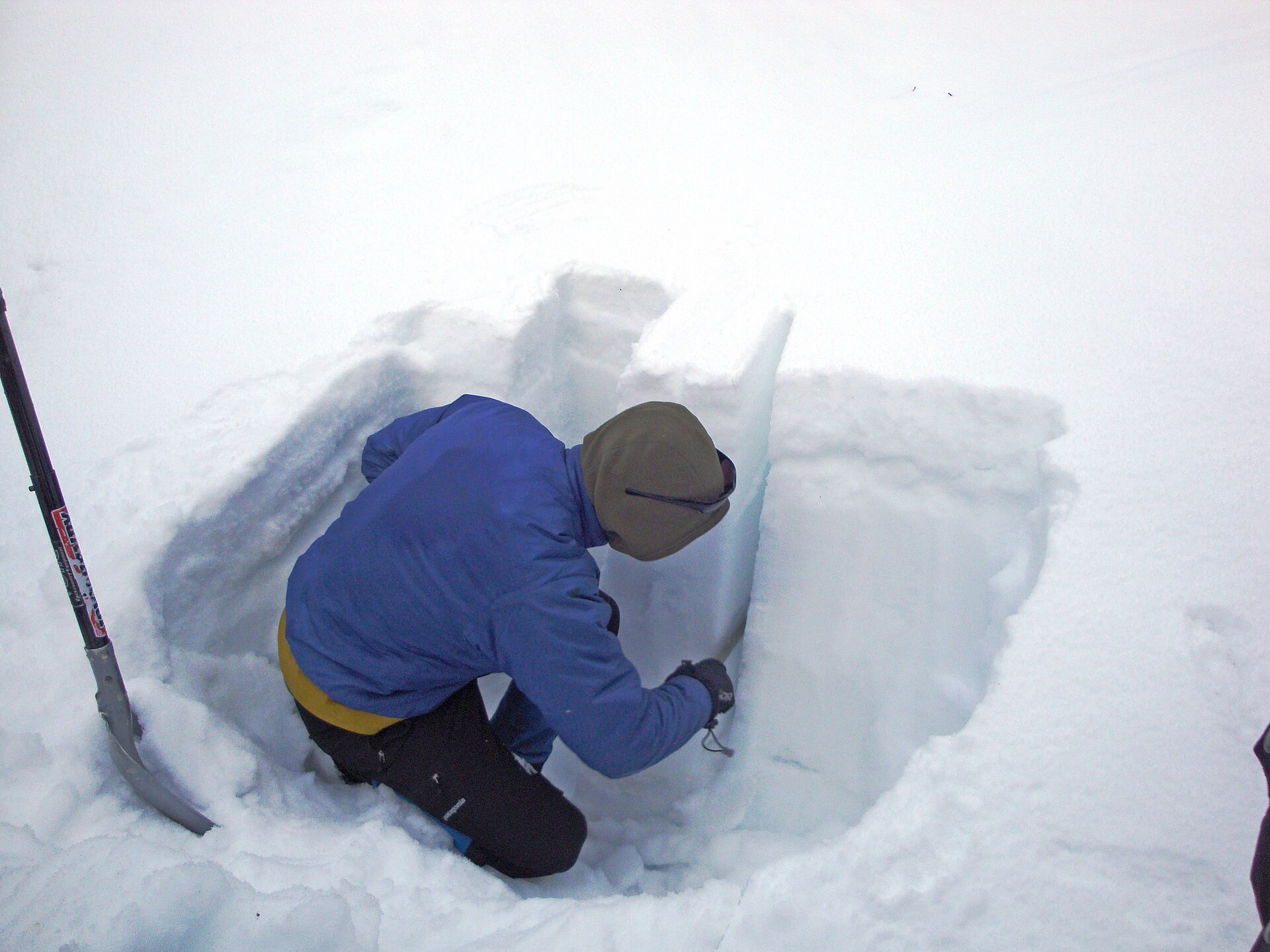 A person kneeling in a snow pit dug down through the snowpack, cutting an isolated column out of the pit wall with a snow saw, a shovel planted in the snow beside them