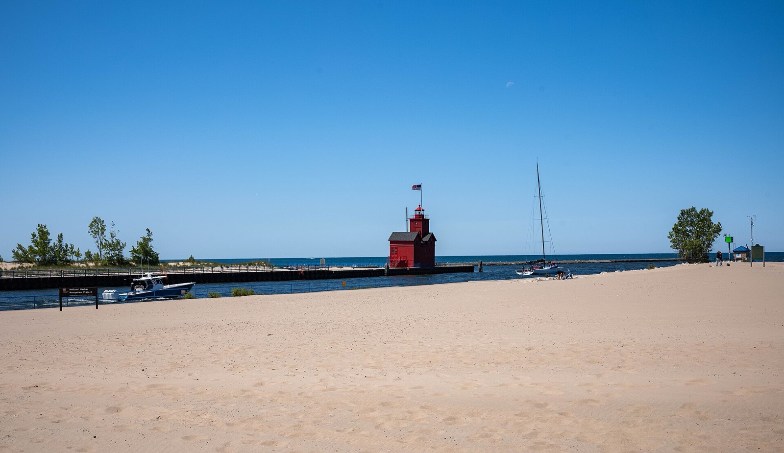 Wide empty sand beach on Lake Michigan under a clear blue sky, with a red lighthouse standing on the channel pier and a sailboat just offshore