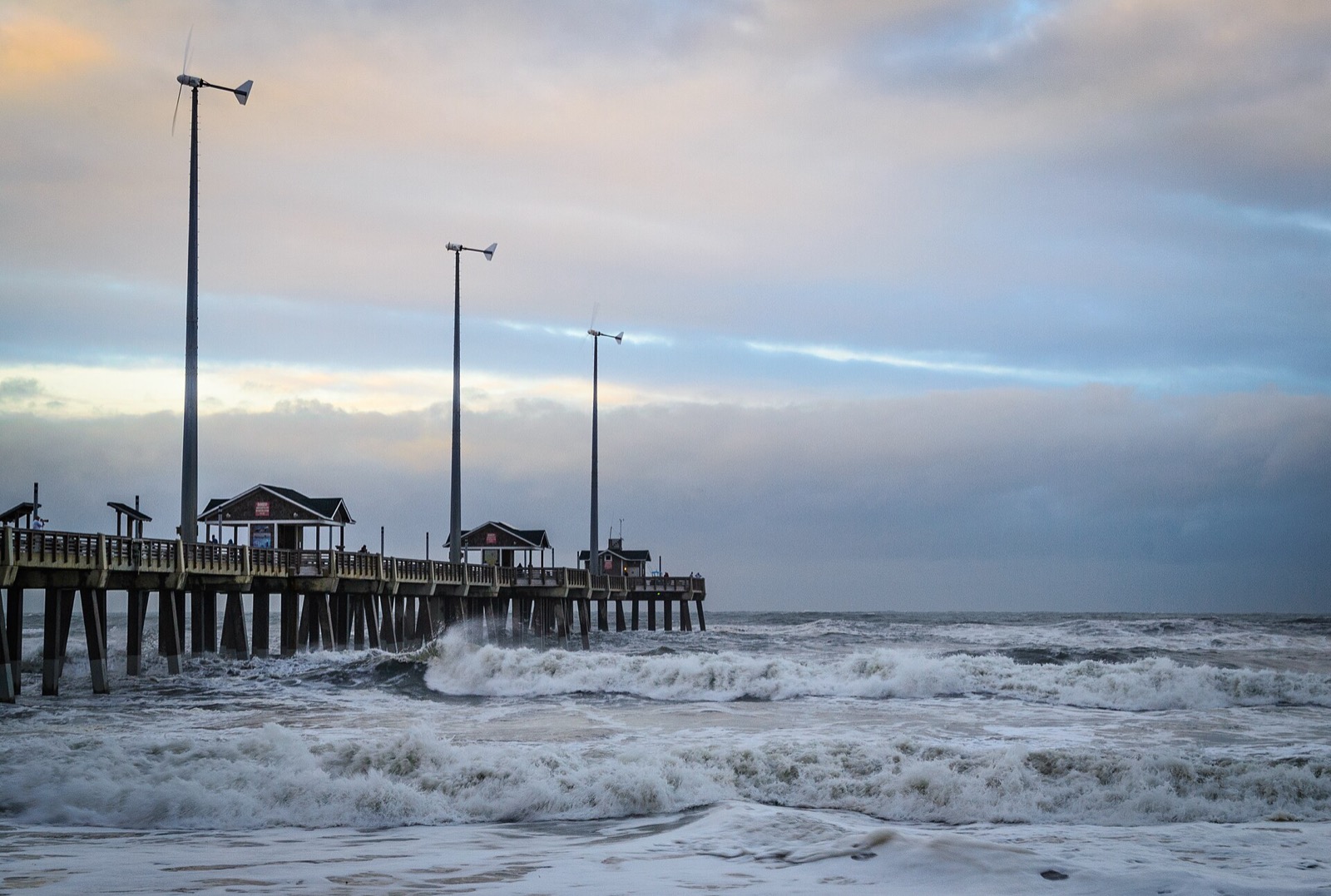 Wooden fishing pier on pilings extending into the ocean at dusk, with heavy surf breaking hard around and through the pilings