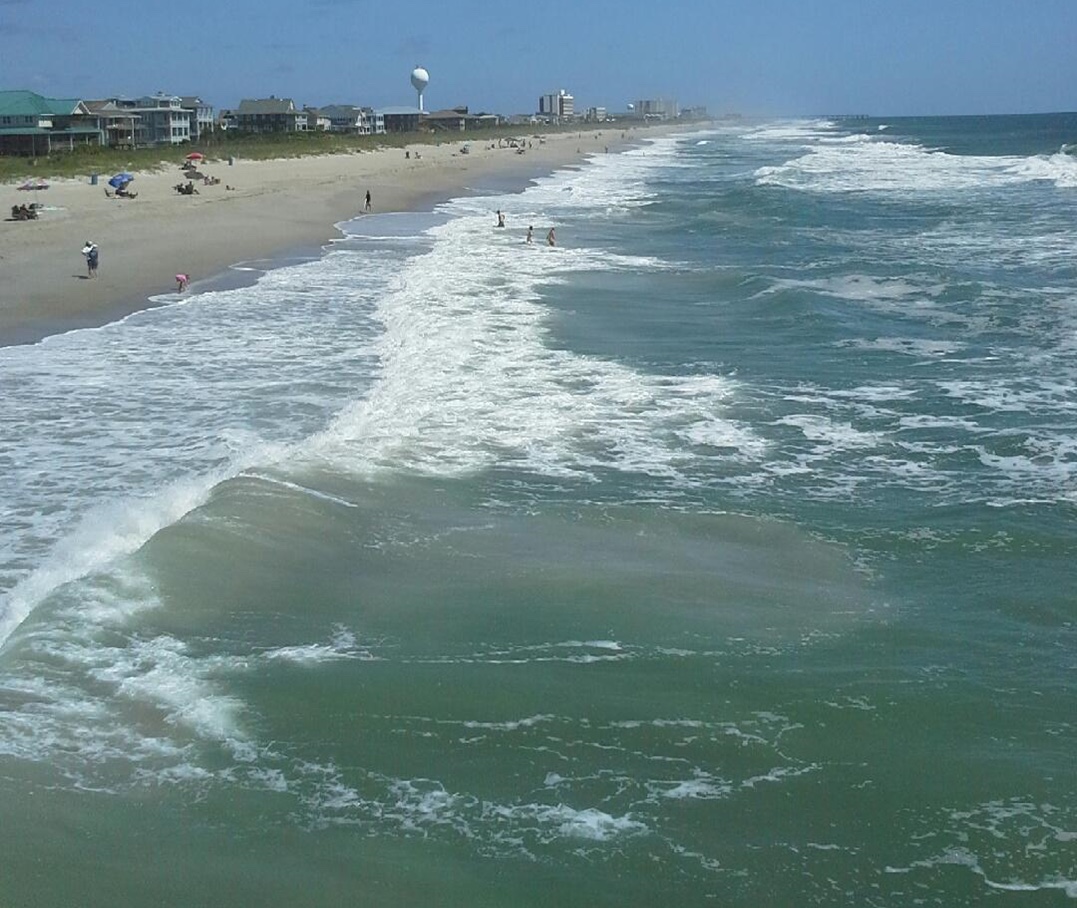 Elevated view along a busy ocean beach showing a rip current, a wide plume of churned, sediment-laden water pushing seaward through a gap in the line of breaking waves