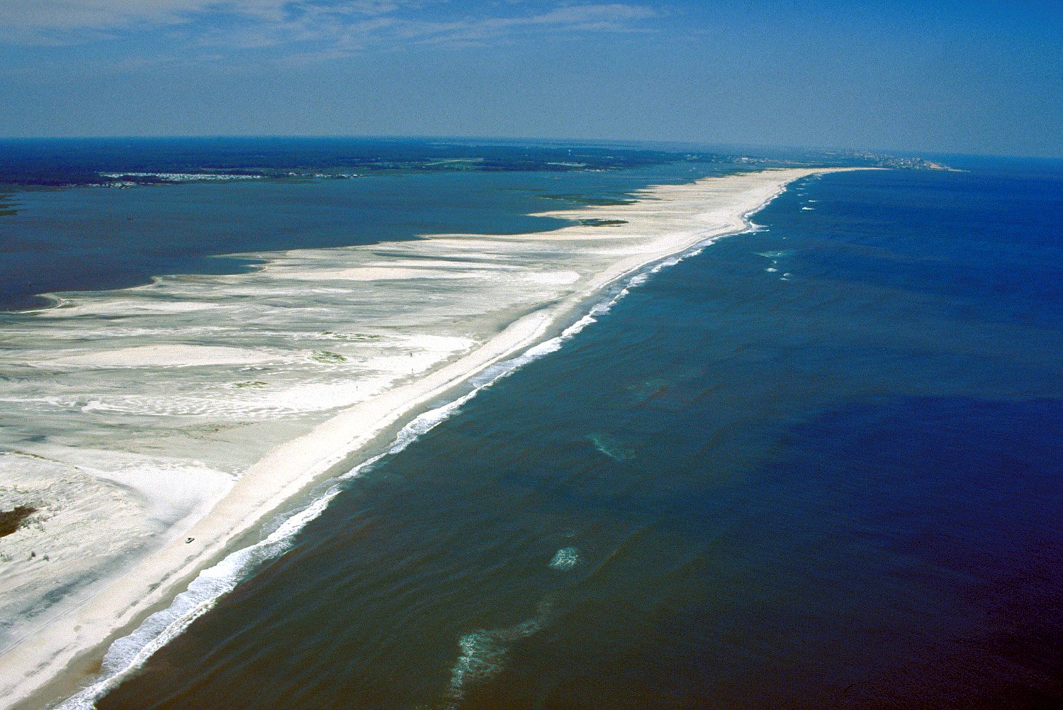 Oblique aerial view along a barrier island beach, with waves breaking in a line offshore and pale submerged sand shoals showing through the shallow water