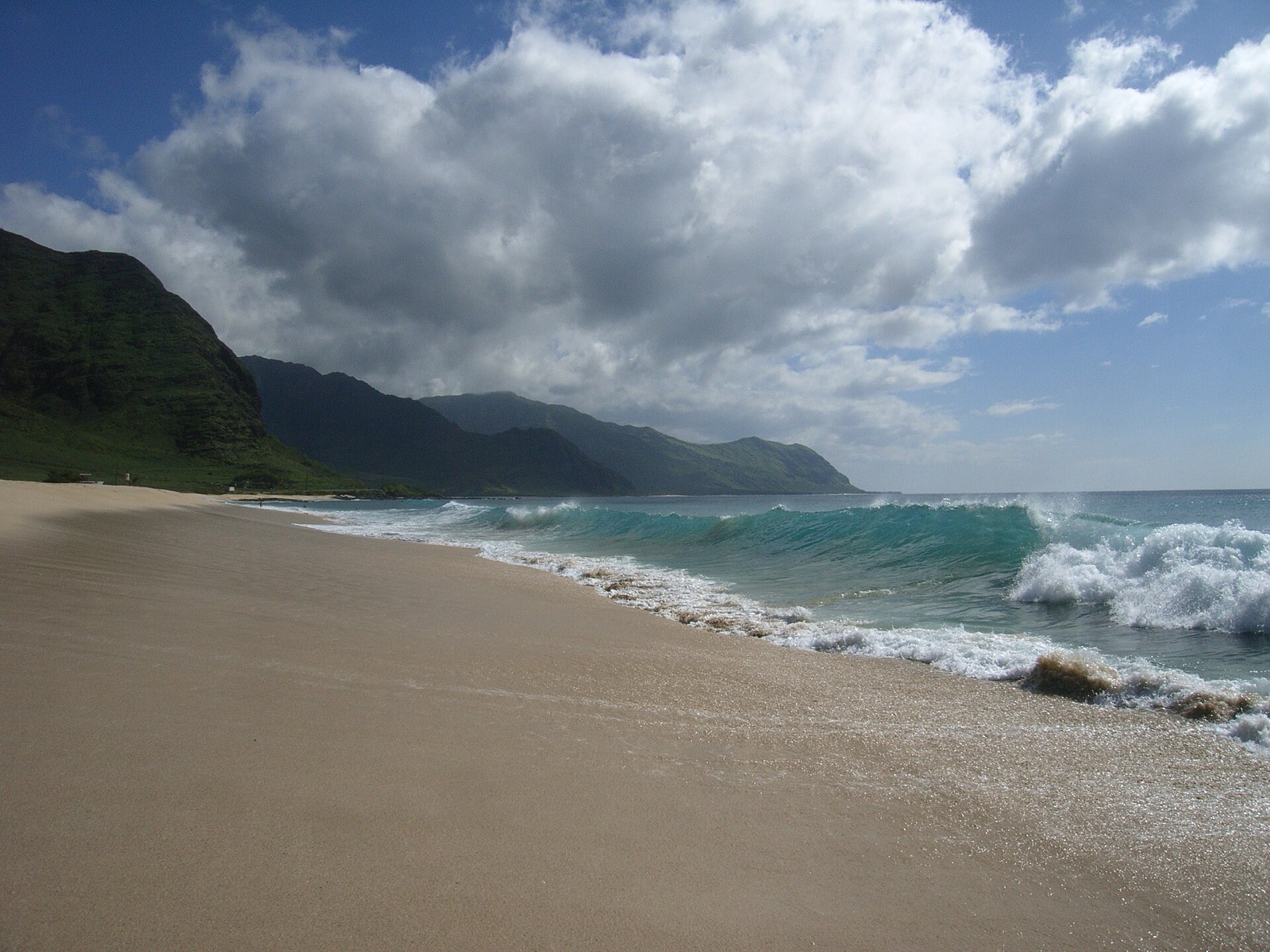 A turquoise wave curling and breaking directly onto the sand at the water's edge of an empty Hawaii beach