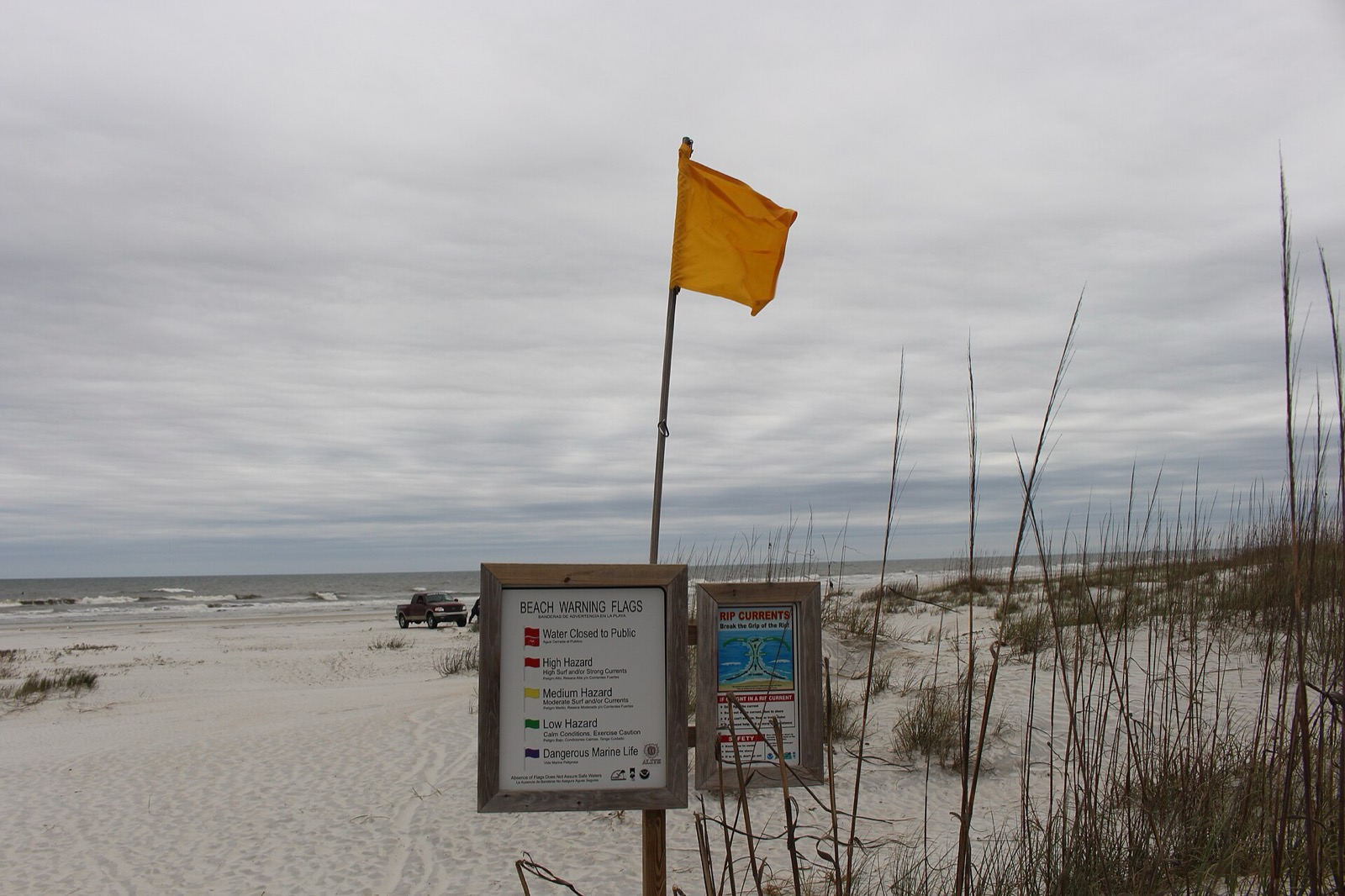Yellow beach warning flag flying from a pole on a Florida beach beside a posted beach warning flag key and a rip current safety sign