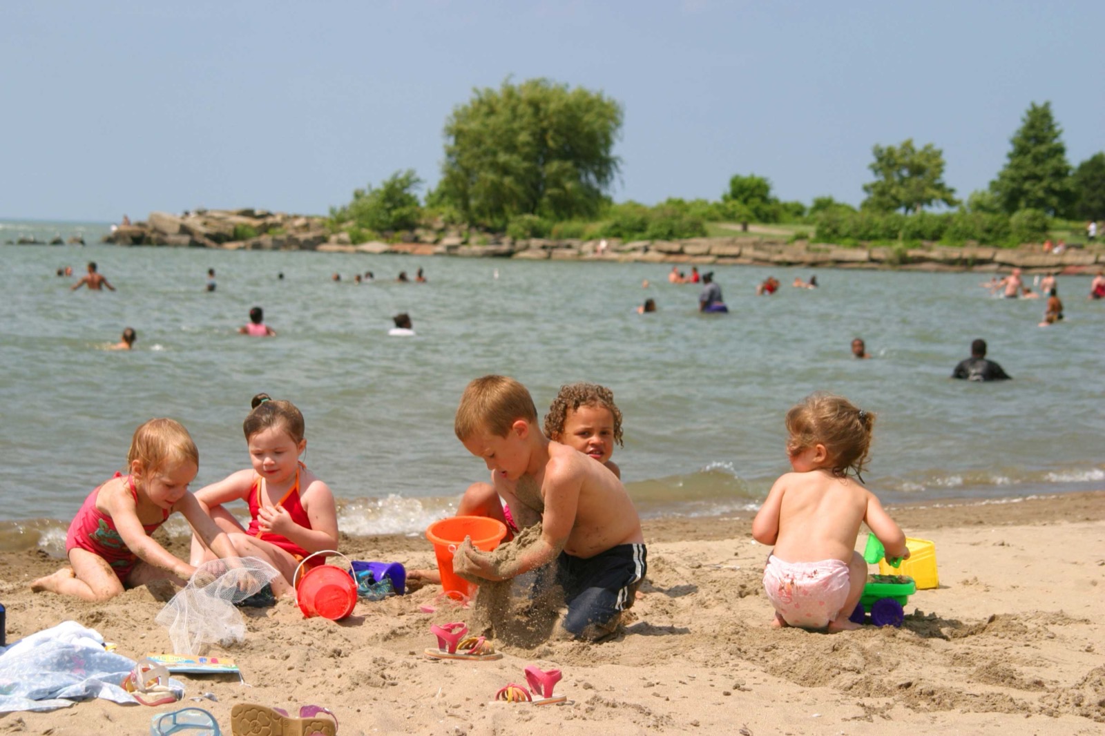 Small children digging in wet sand at the edge of a lake while swimmers stand in the shallow water behind them under a hazy summer sky