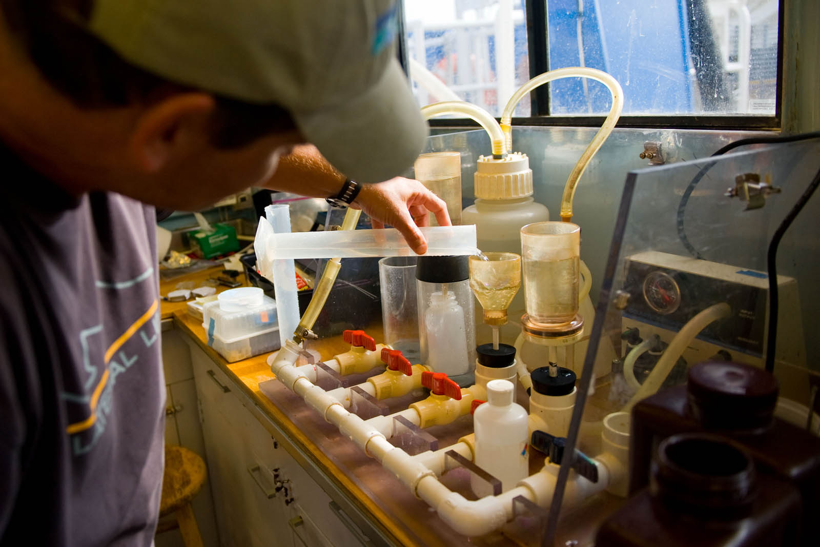 A technician pouring a water sample into a filtration funnel on a bench manifold of clear cylinders and valves
