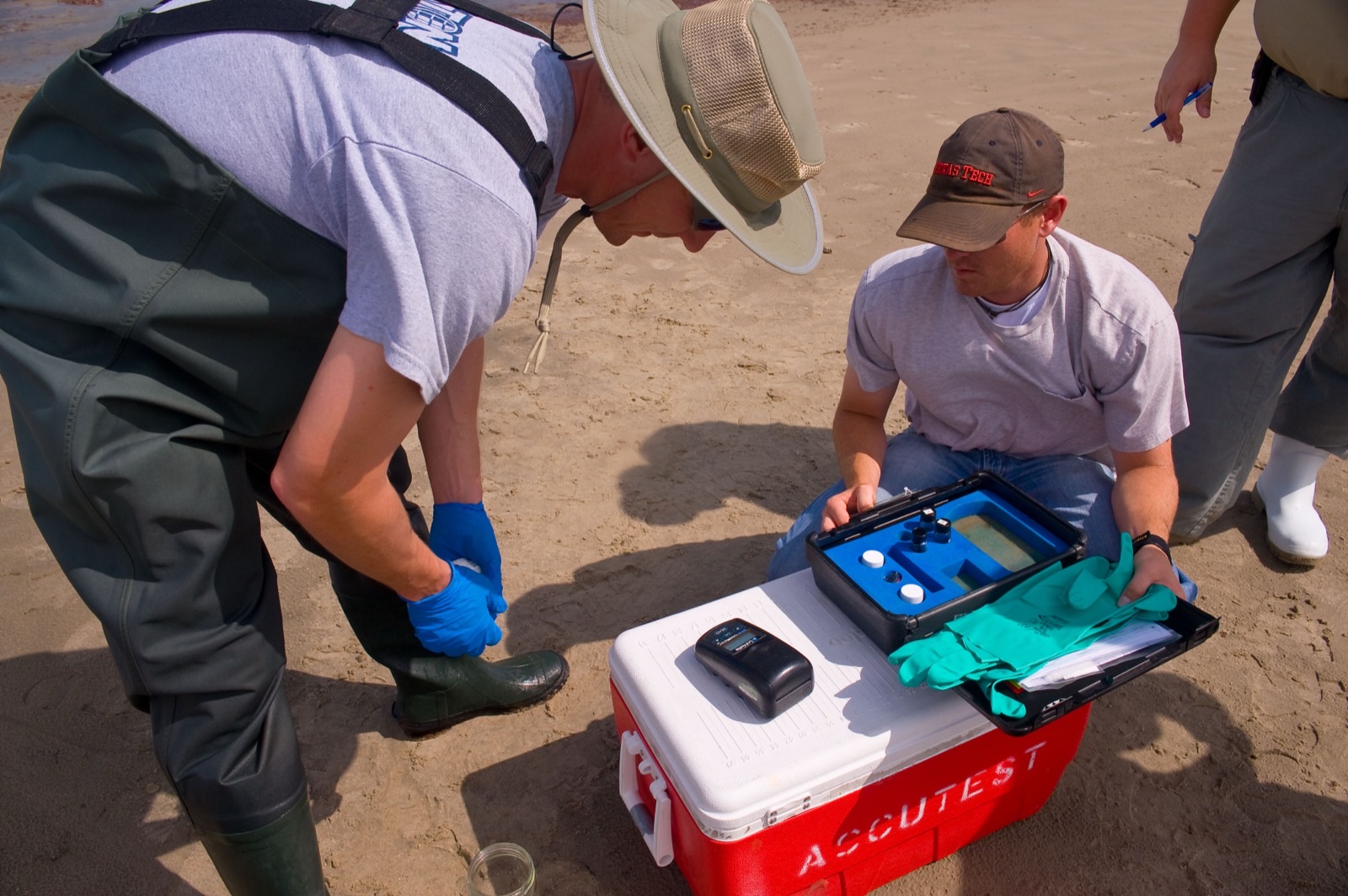 Two people crouched on wet sand beside a red cooler and a portable test kit, one in chest waders holding a sample in a gloved hand