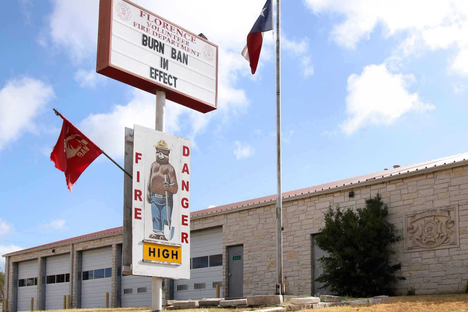 A volunteer fire department letter board reading BURN BAN IN EFFECT above a Smokey Bear fire danger sign showing HIGH, with a red flag flying beside it
