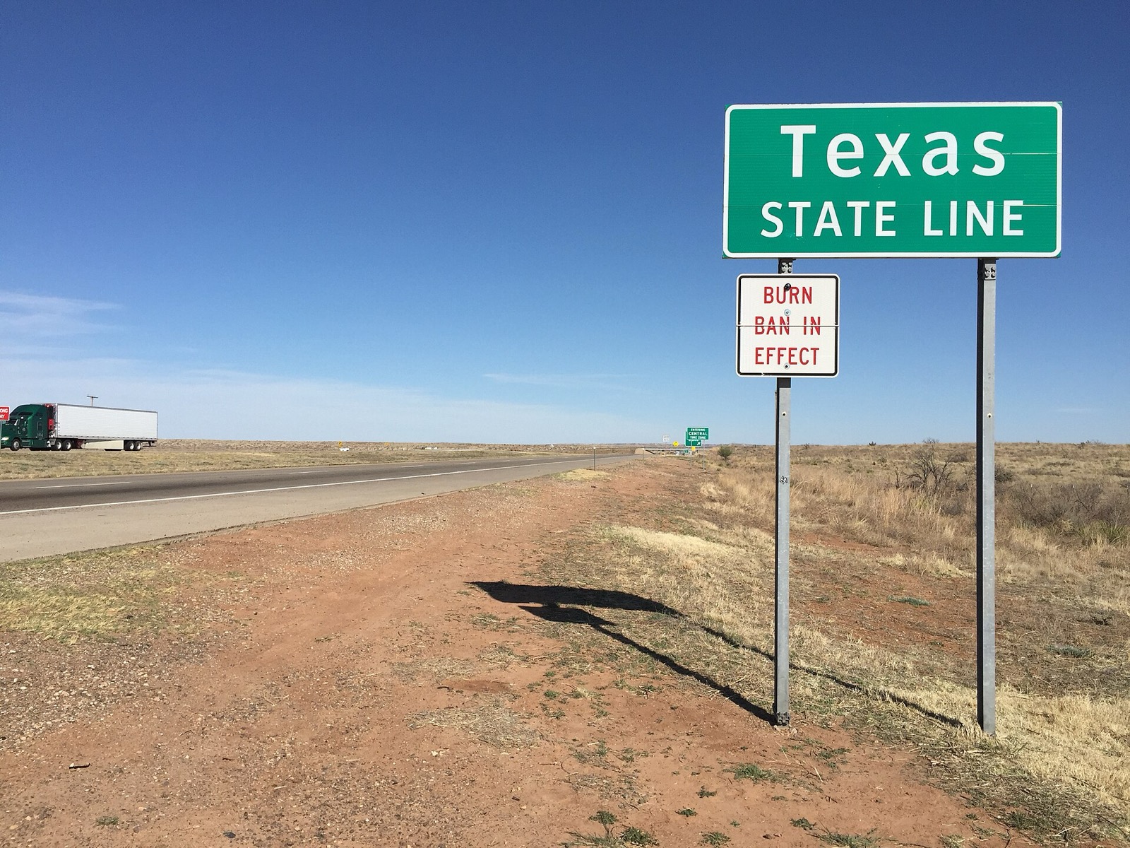 A green Texas State Line sign beside an interstate in open country, with a small white sign bolted under it reading BURN BAN IN EFFECT