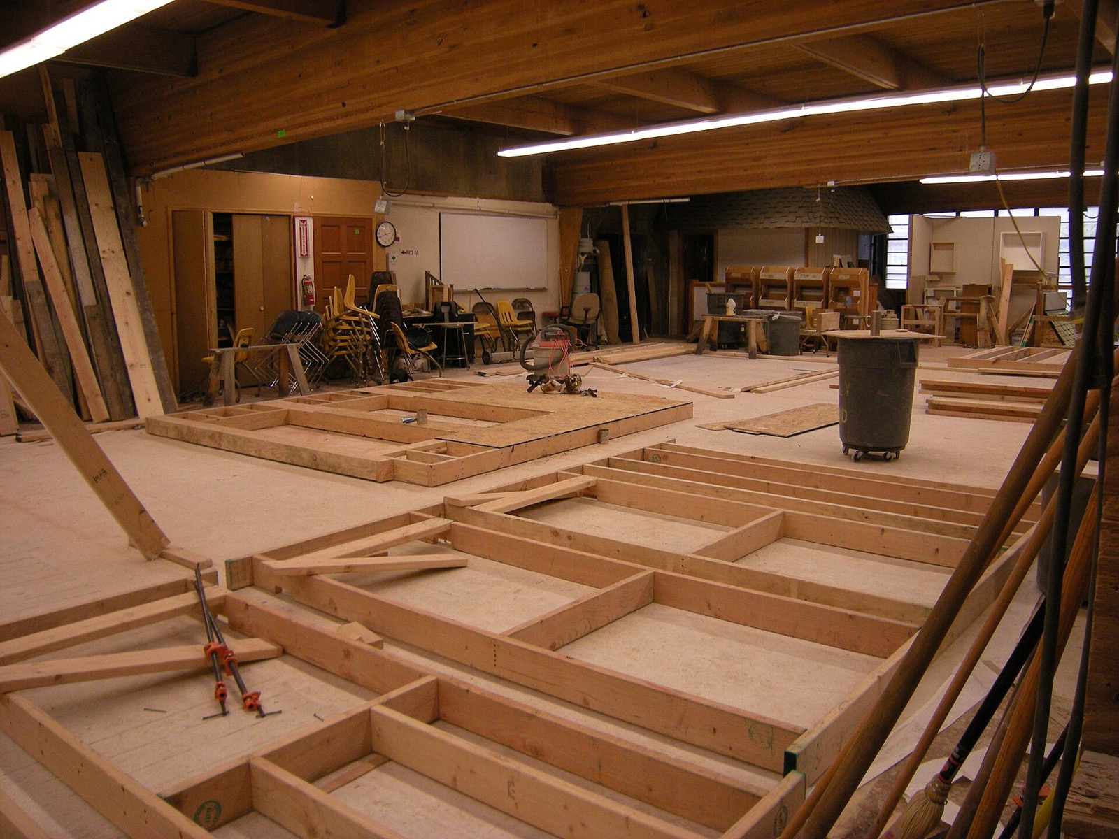 Stud walls for a house laid out flat on a shop floor, framed in dimensional lumber with clamps and offcuts around them.