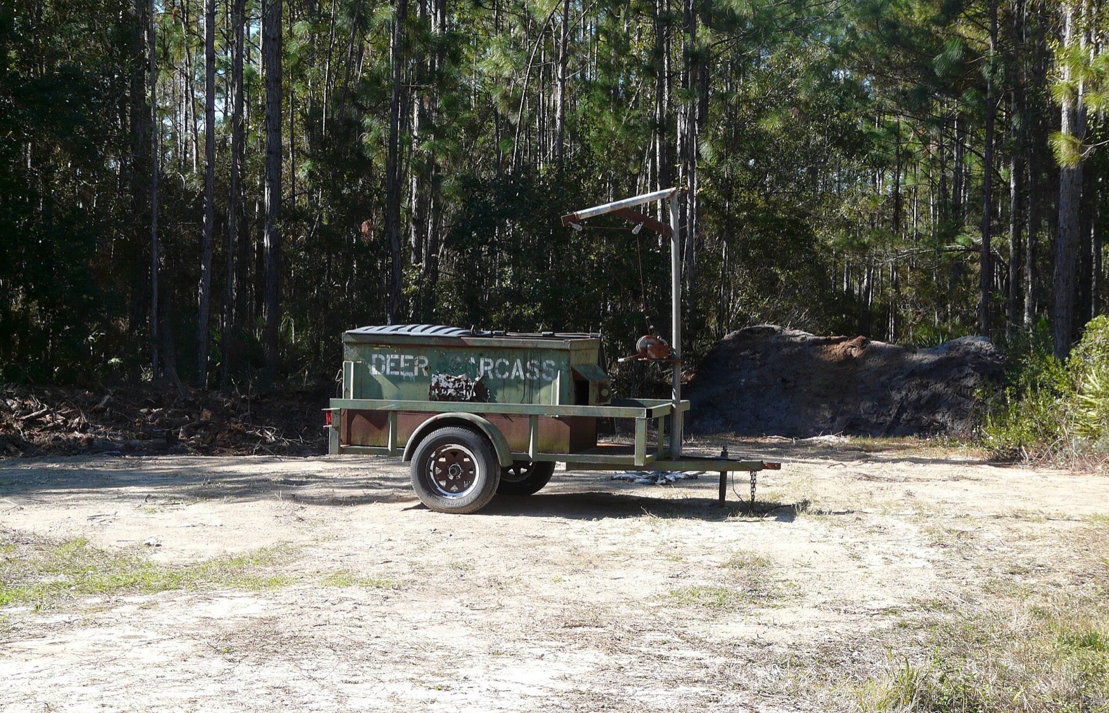 A green steel dumpster stencilled DEER CARCASS mounted on a single axle trailer in a sandy pine clearing, with a hoist gantry beside it and a pile of soil behind