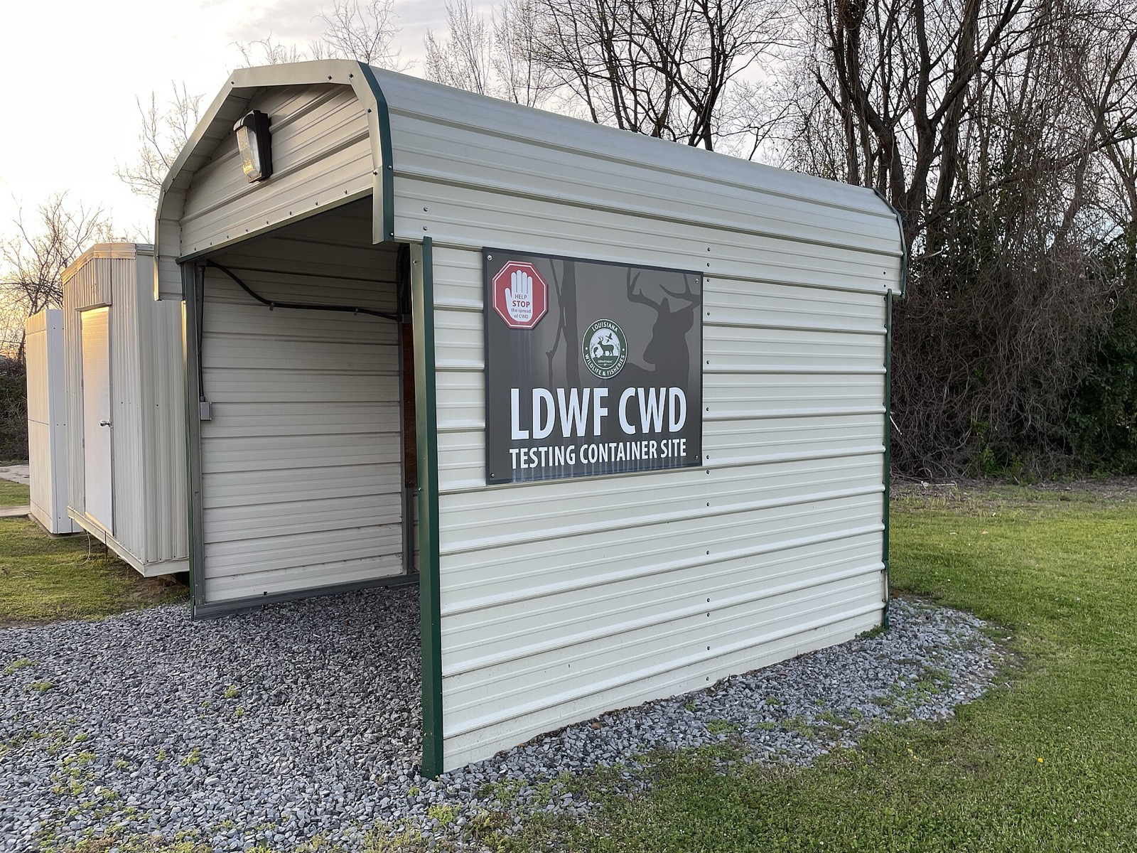 A small white steel shelter on a gravel pad with one open bay and a sign reading LDWF CWD TESTING CONTAINER SITE beside a deer silhouette, bare trees behind it at dusk