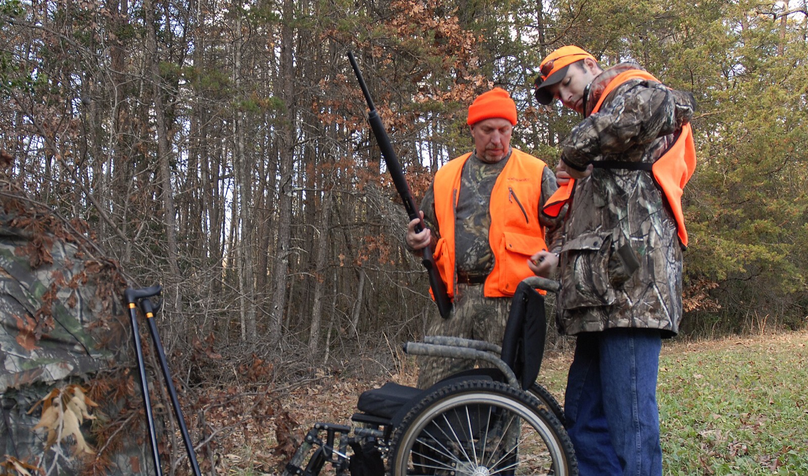 Two hunters in blaze orange and camouflage beside a wheelchair at the edge of bare winter woods, one helping the other with gear
