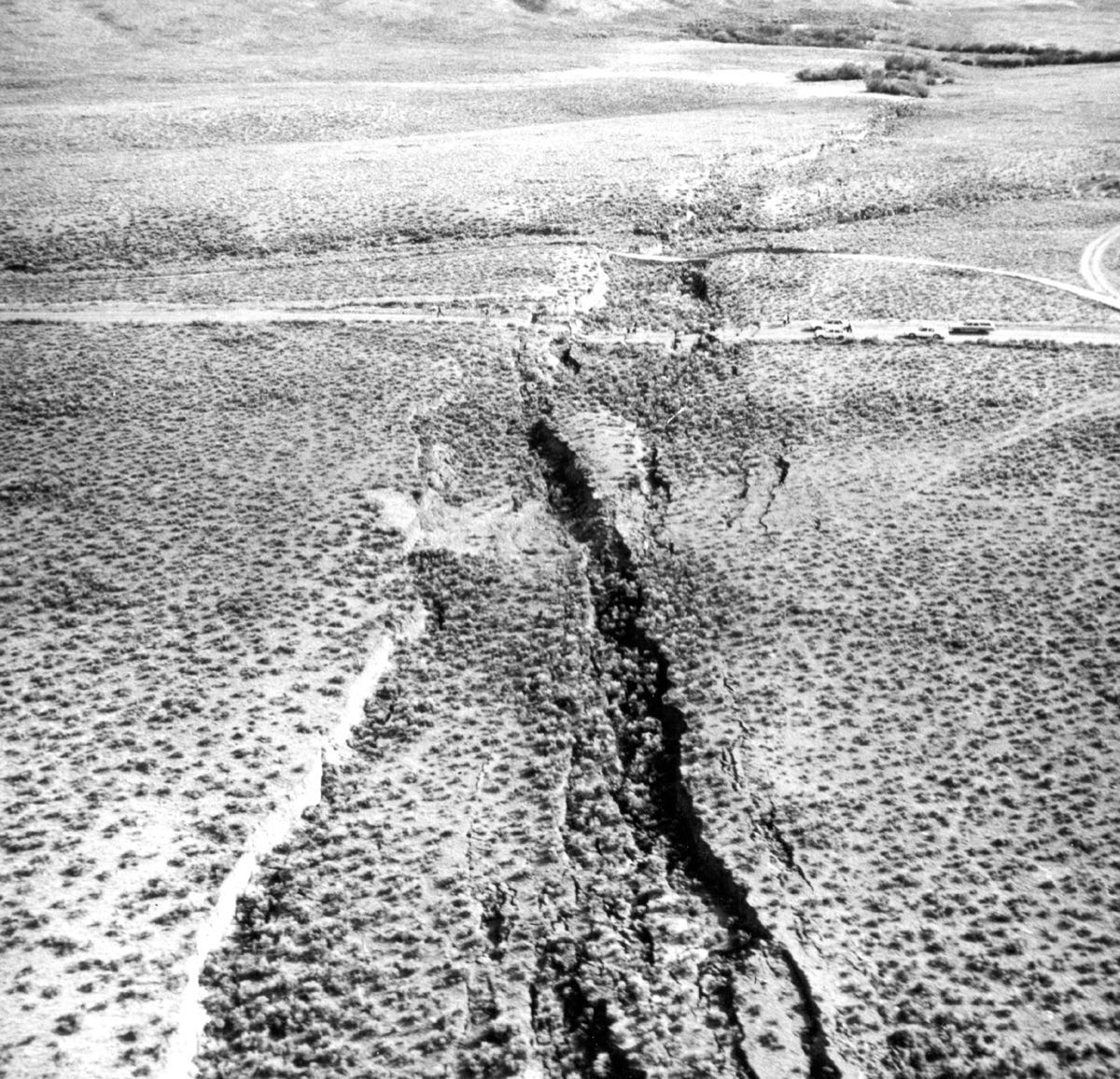 Black and white aerial photograph of fresh fault scarps cutting across a sagebrush valley floor at the foot of a mountain range