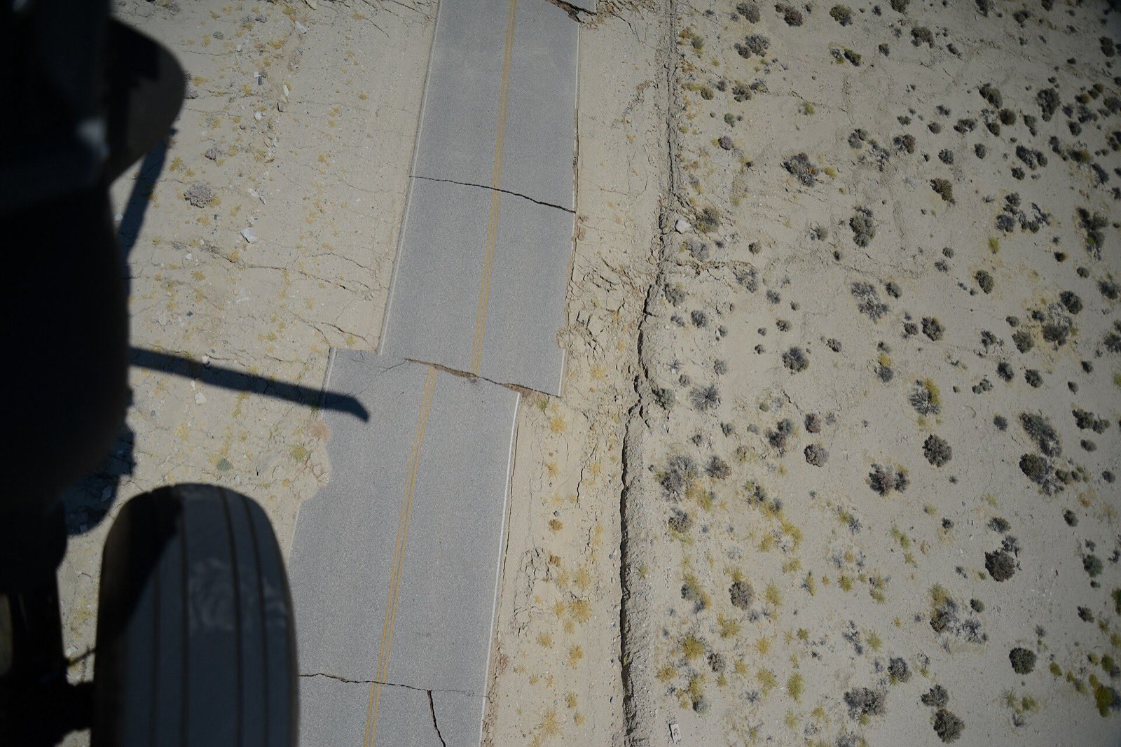 A two lane desert road broken and shifted sideways where a fault rupture crosses it, seen from a helicopter with people standing beside the break