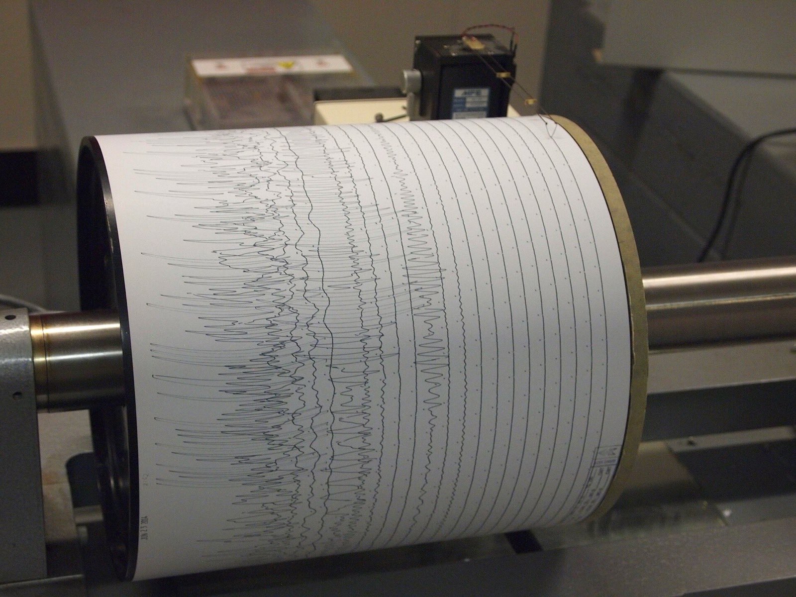 A seismograph drum wrapped in paper with rows of ink traces, the pen drawing a new line as the drum turns