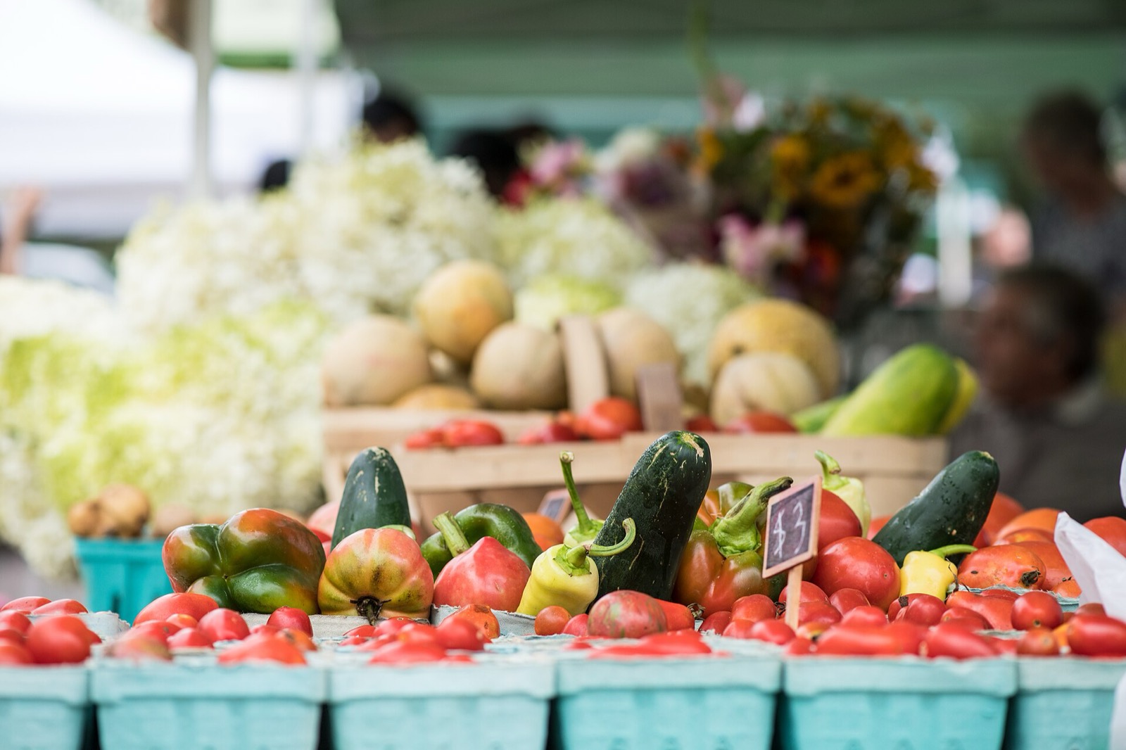 Tomatoes, peppers and cucumbers on a market table in turquoise pulp baskets, with melons and cut flowers behind
