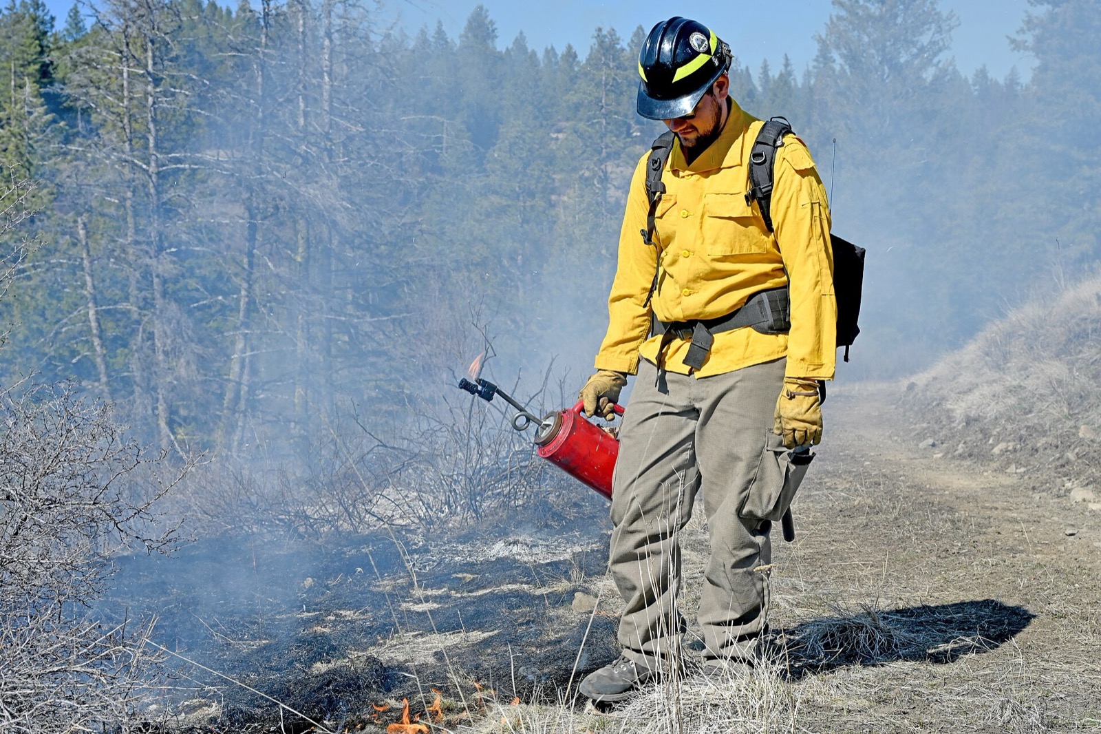 A firefighter in a yellow shirt and hard hat tipping a red drip torch along the edge of a dry hillside, low flames and pale smoke in the brush beside him