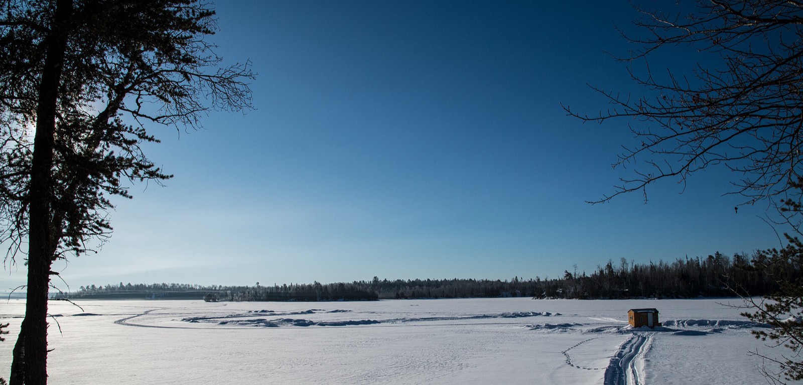 A wide view from a wooded shore across a snow covered frozen lake in low winter sun, a single small yellow ice shanty far out on the ice at the right with one set of tracks running out to it, and a dark treeline on the far shore