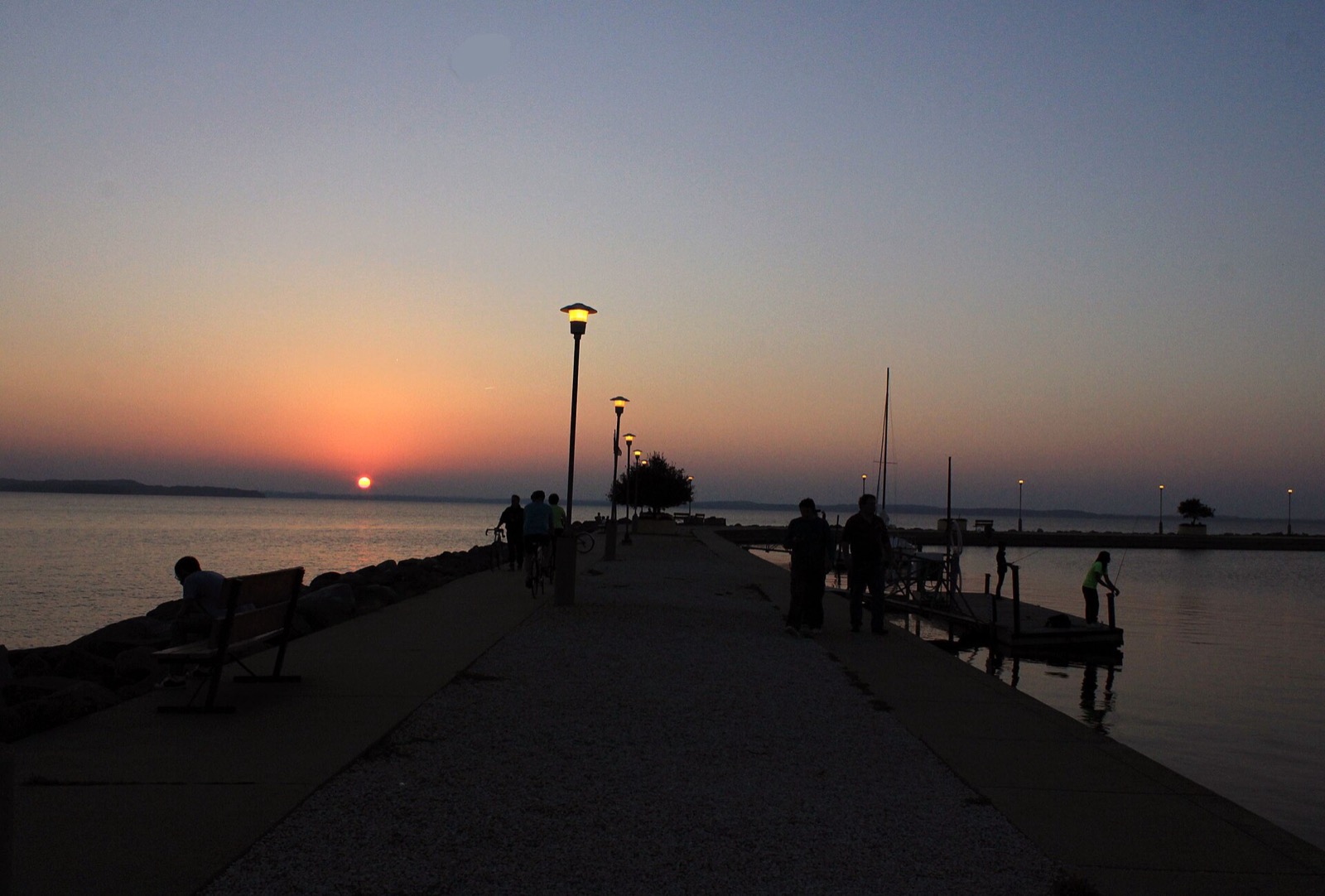 People fishing and walking on a long concrete pier at sunset, the sun a small orange disc on the horizon over a lake