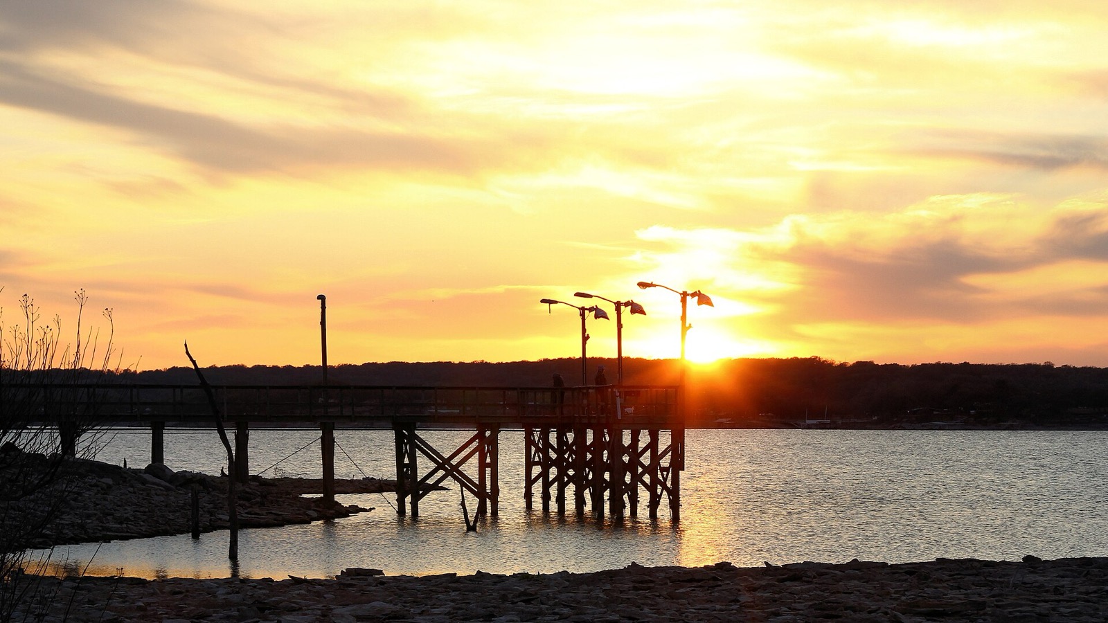The silhouette of a wooden fishing pier with lamp posts reaching into a lake, the sun setting behind it under a gold sky