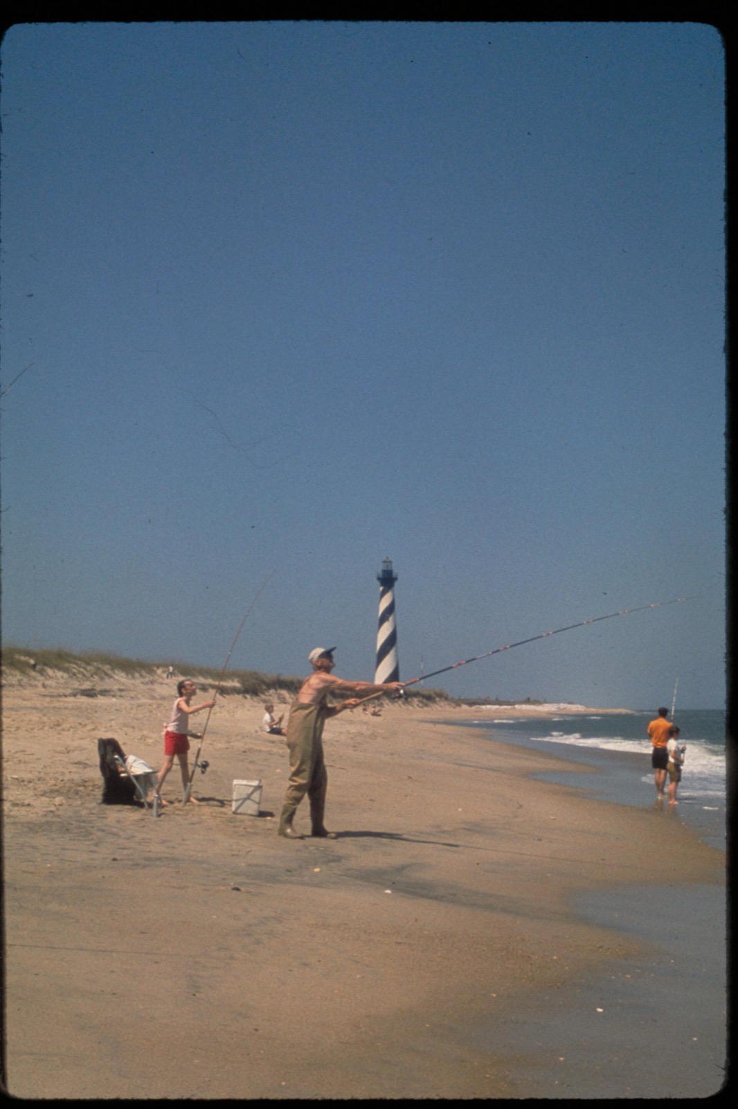 Surf anglers casting from a sandy beach under a clear blue sky, with the black and white spiral of the Cape Hatteras lighthouse behind them