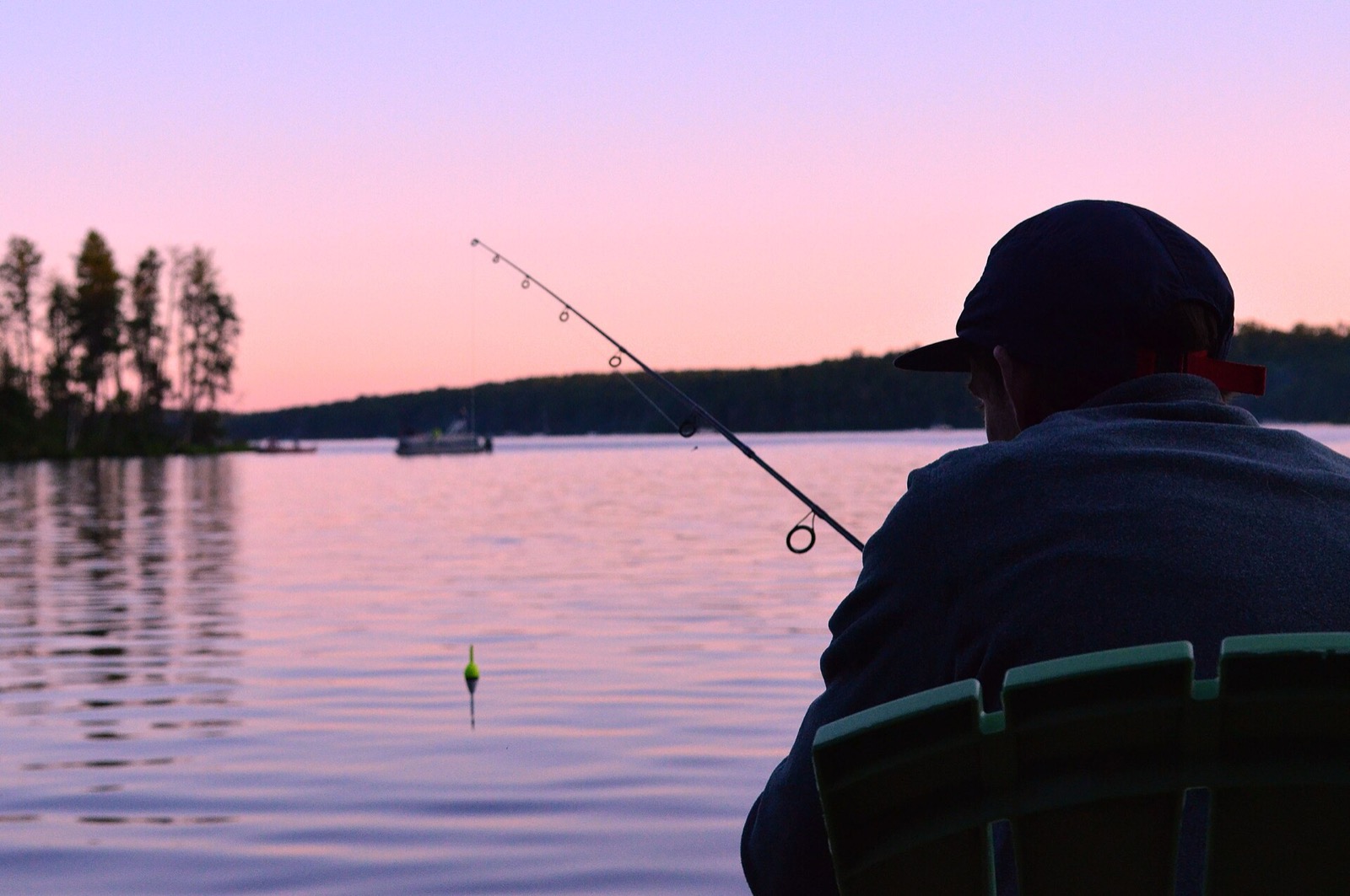 An angler in a cap seen from behind, holding a rod over a calm lake at dusk, with a bobber on pink water and a pine shore beyond