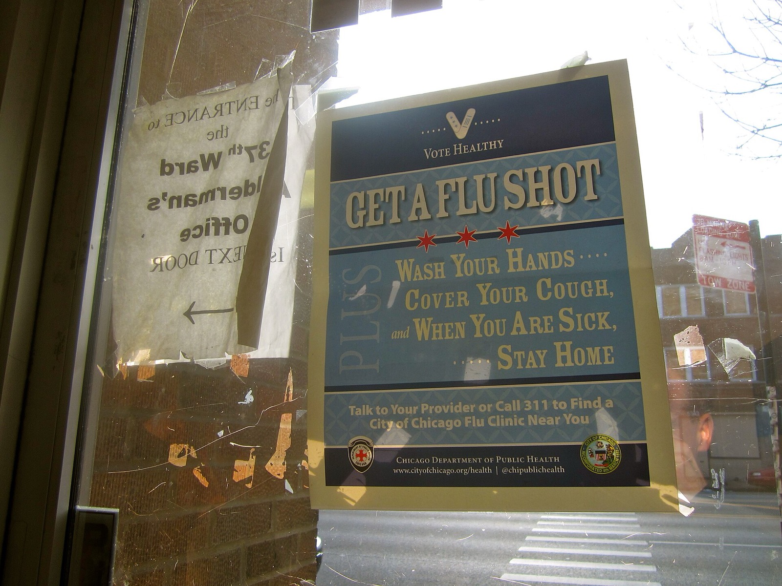 A Chicago Department of Public Health poster taped inside a glass door reading Get a Flu Shot, Wash Your Hands, Cover Your Cough, and When You Are Sick, Stay Home