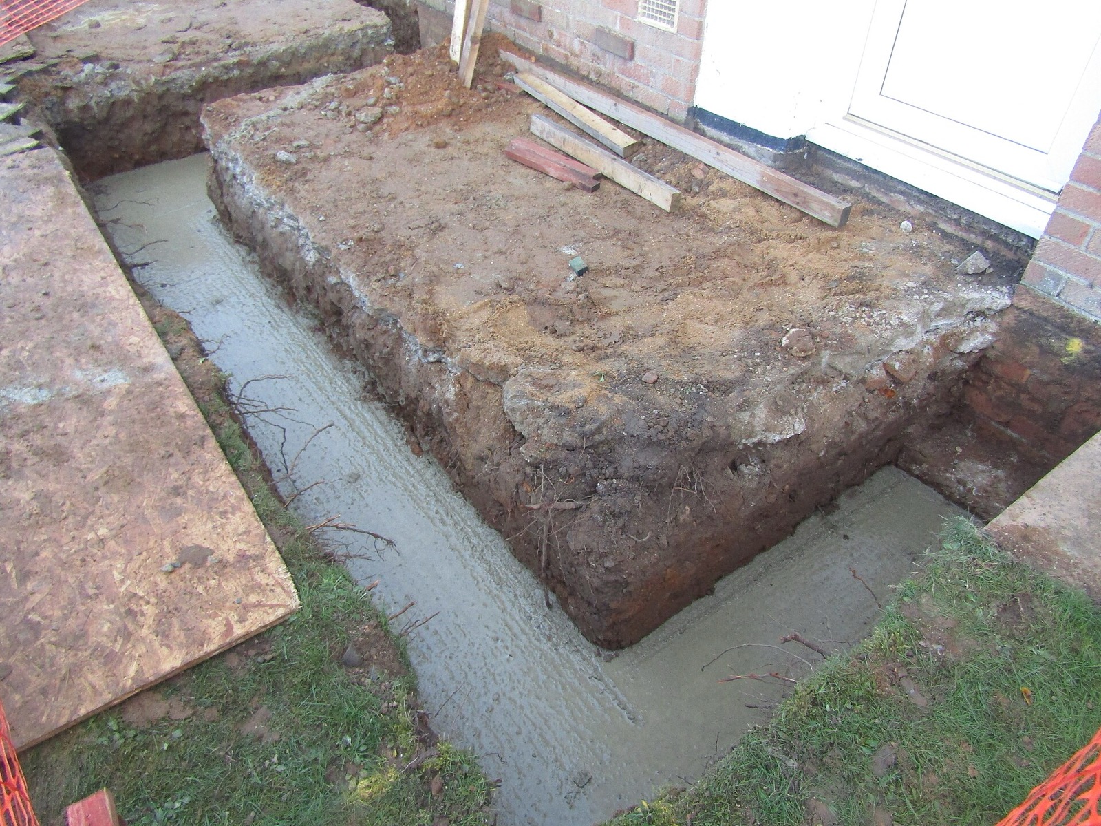 An open footing trench cut around three sides of a rectangle of undisturbed earth beside a brick house, wet concrete lying in the bottom of the trench