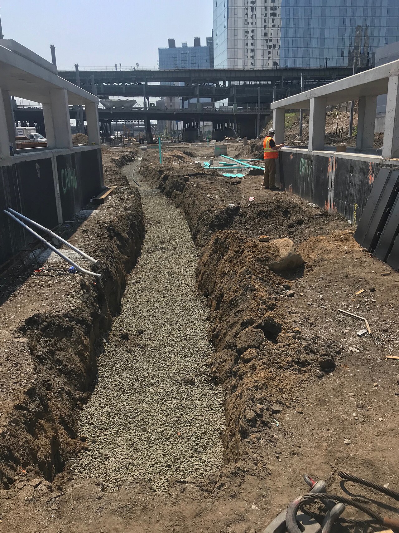 A long open trench with a gravel bed running down the middle of a city construction site, lengths of blue water pipe staged on the ground beside it