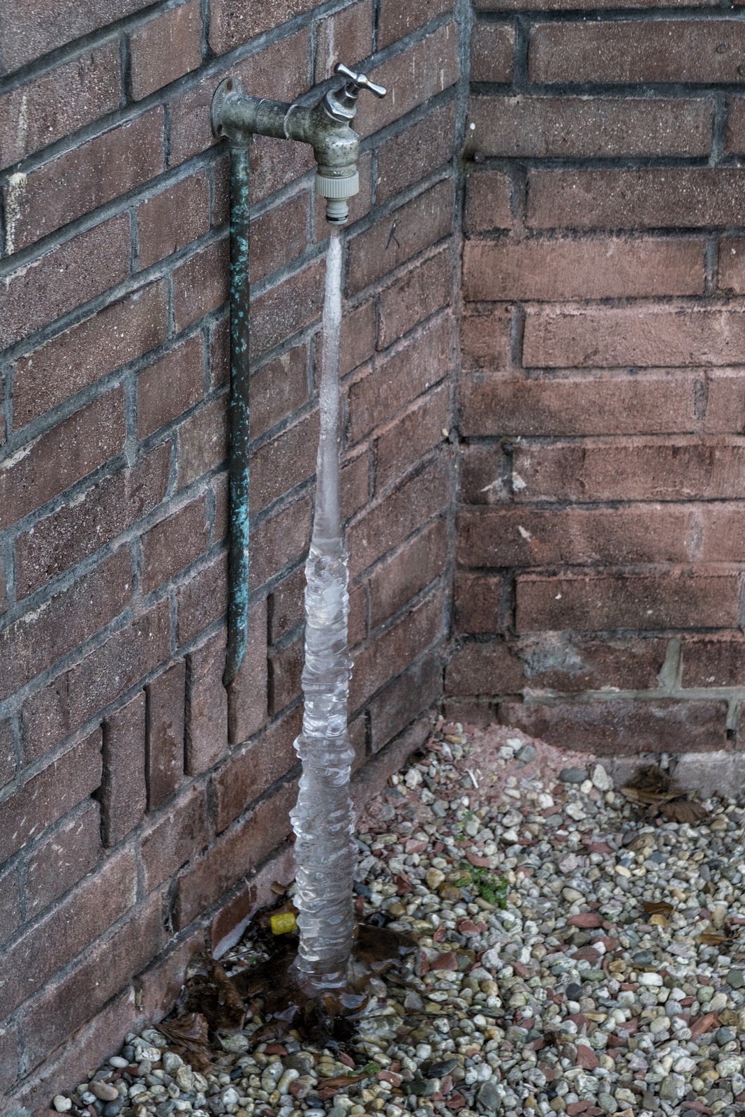A solid ribbed column of ice hanging from an outdoor tap on a brick wall down to the gravel below, with a copper supply line running down the brickwork