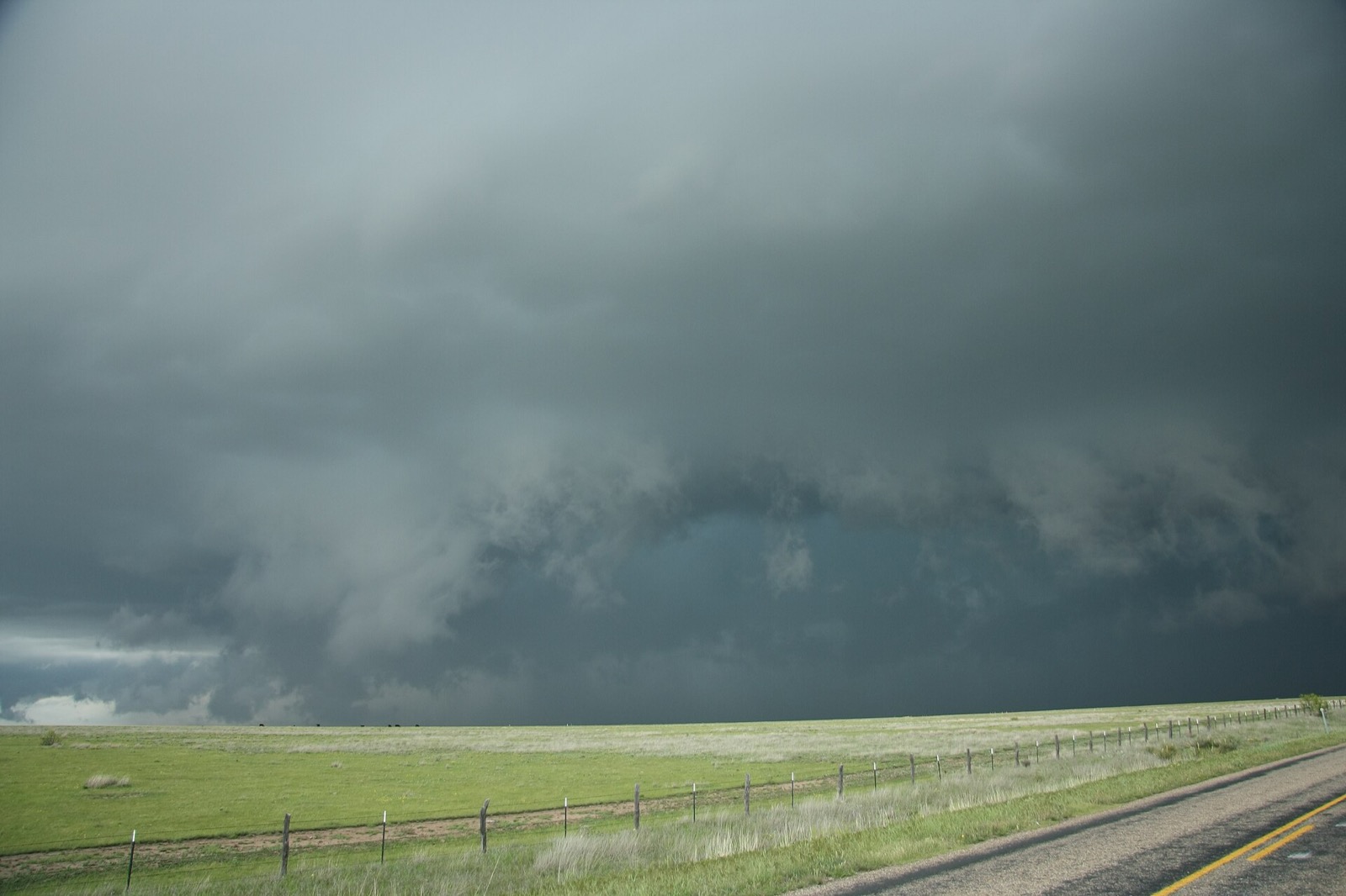 A dark rotating wall cloud hanging below a severe storm base over open green pasture, with a barbed wire fence line and a two lane road in the foreground