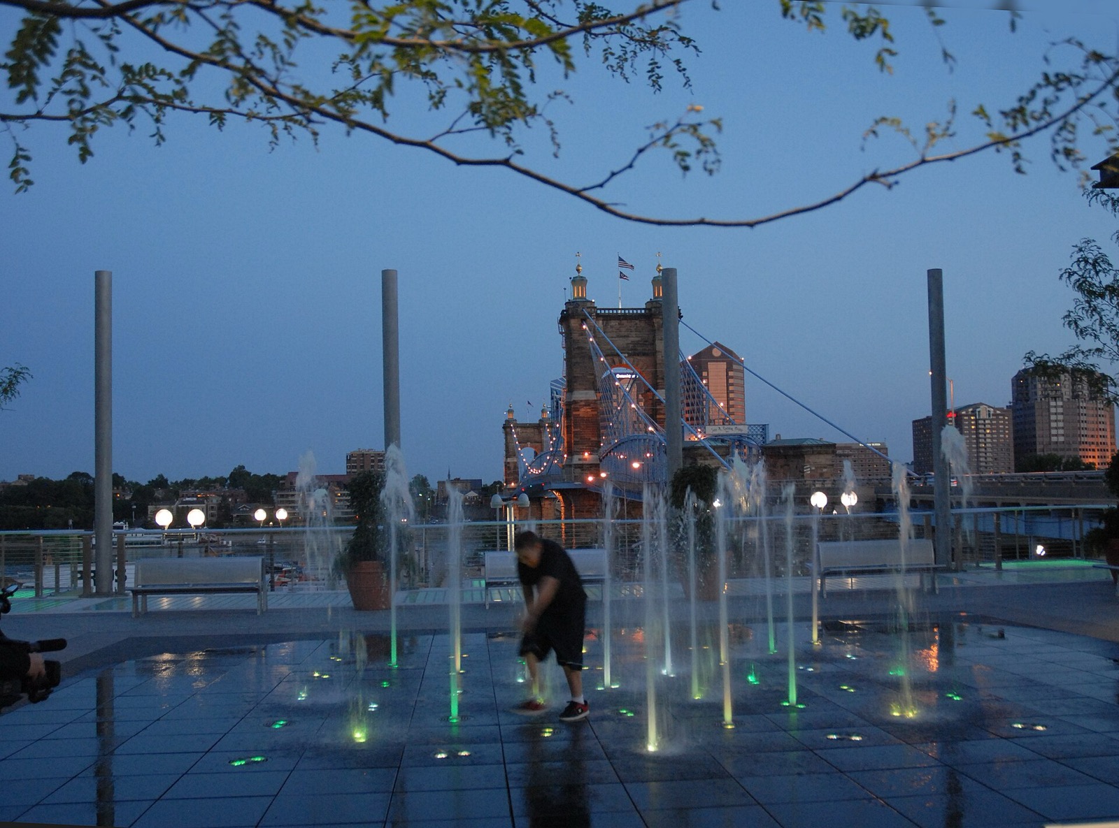 People still out at a lit riverfront fountain in Cincinnati at dusk, with the Roebling Bridge and downtown skyline behind them