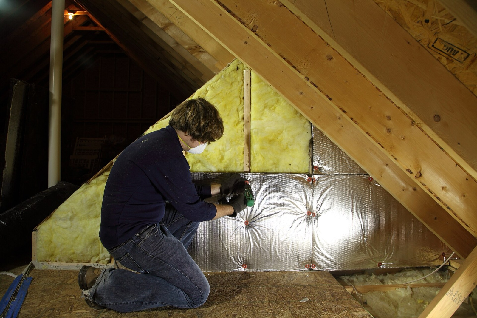 An installer in a dust mask and gloves kneeling on joists in a bare attic, laying a fresh batt of pink insulation into the bay beside several already filled, with the roof sheathing close overhead