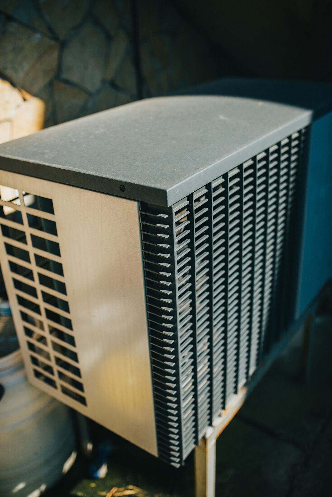A tall grey outdoor condenser unit standing on a frame against a stone garden wall, its fins and louvred side panel filling the frame, no maker’s plate or badge visible anywhere on it