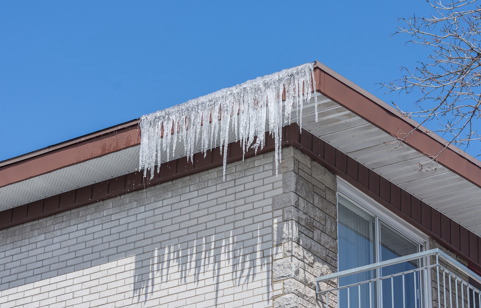 Icicles hanging in a row from the eaves of a low brick apartment building against a clear blue sky, with a balcony rail at the right edge