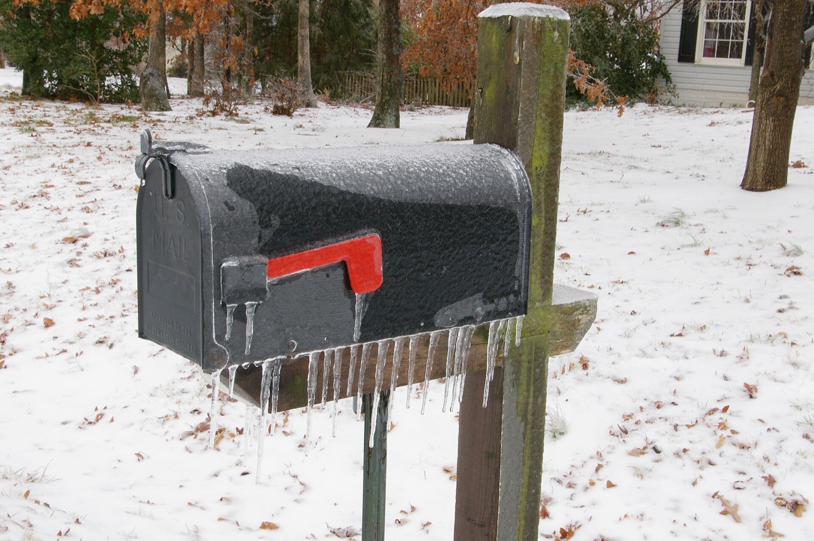 A black US mailbox with its red flag up, glazed in ice with icicles hanging from its underside, standing on a wooden post in a snow-covered yard