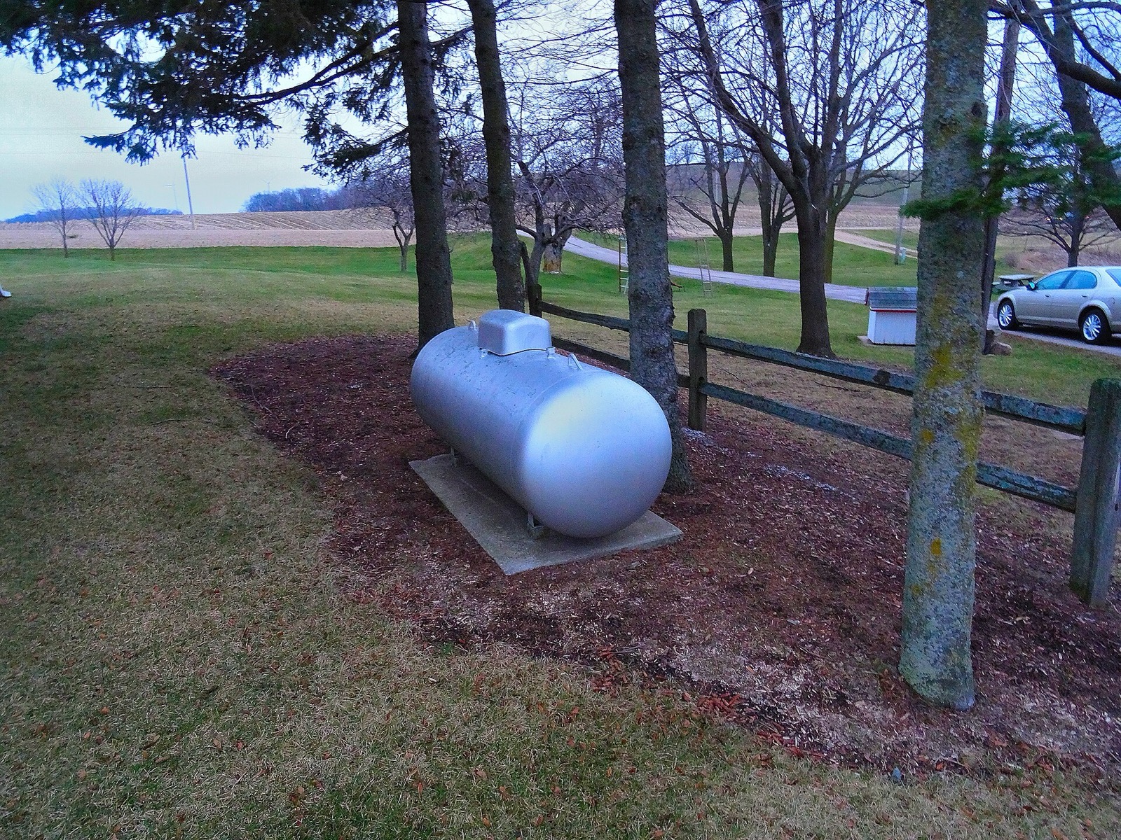 A white residential propane tank standing on a concrete pad beside a mown yard, with bare trees and a wooded ridge behind it