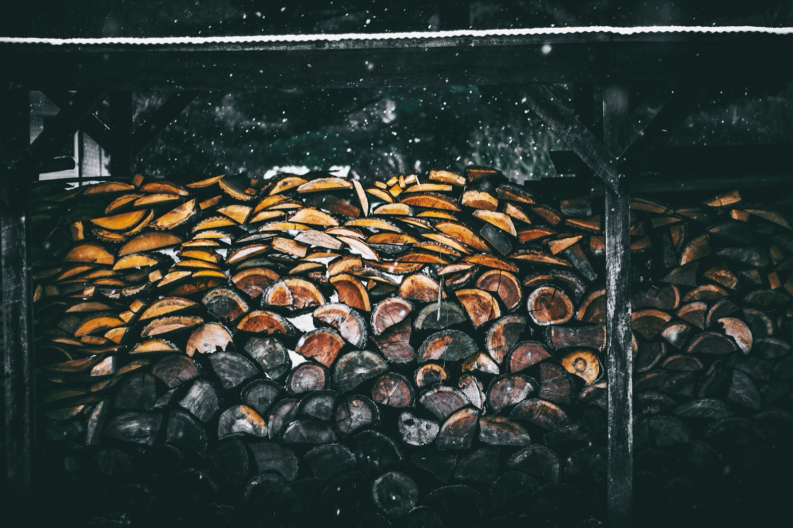 Split firewood stacked under an open sided wooden shed while snow falls in the dark behind it