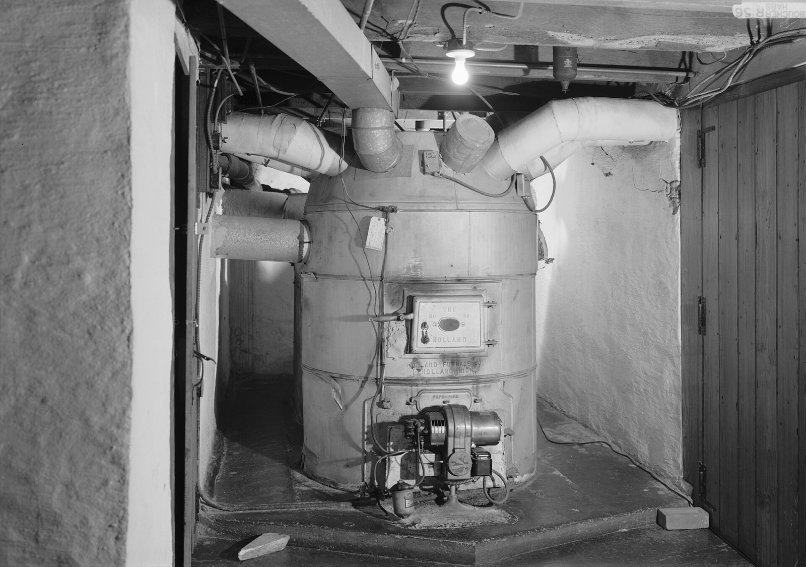 A round cellar furnace with wide ducts branching from its top like arms, an oil burner bolted to its base, lit by a single bulb