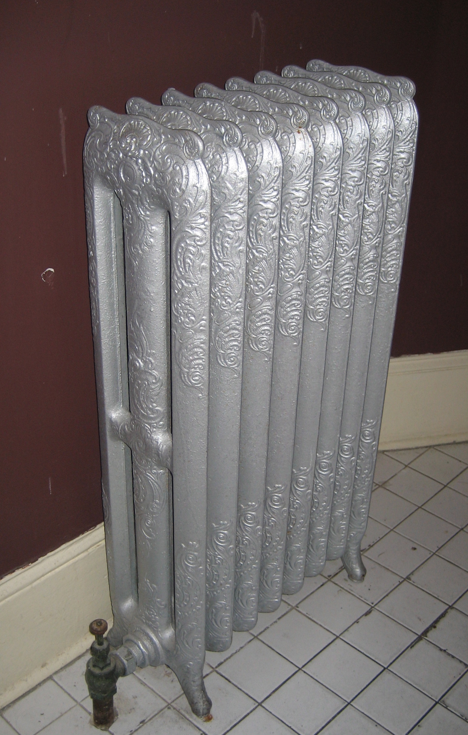 An ornate cast iron column radiator painted silver, standing against a dark red wall on a tiled floor