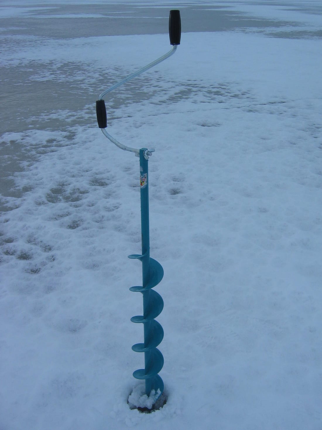 A turquoise hand auger with a cranked steel handle standing upright in a freshly cut hole, its spiral blade about ten inches wide, on a wind scoured snow surface