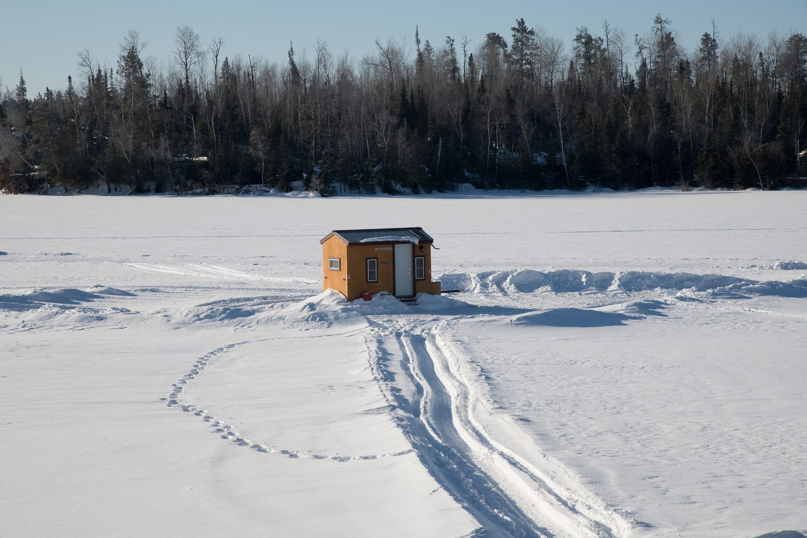 A single small orange plywood ice fishing house with a white door standing alone on a wide snow covered lake, a strip of dark conifer forest behind it, and a snowmobile track curving away across the snow in front