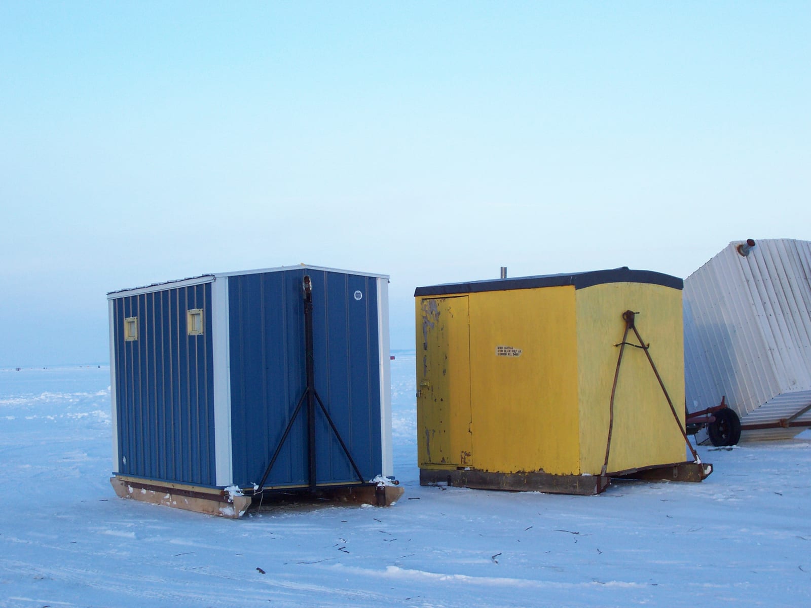 Three ice fishing shanties standing on packed snow at dusk, a blue metal one and a yellow wooden one on timber skids with tow bars folded down, and a third silver one on a wheeled trailer behind