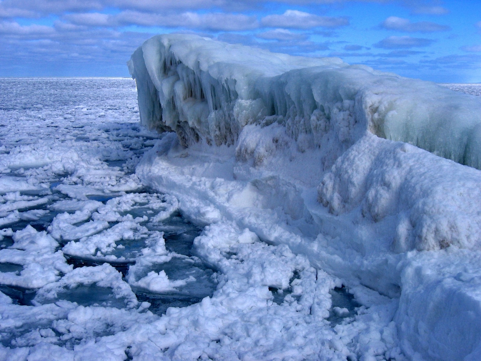A wall of shelf ice rising above a lake surface strewn with broken plates of new ice and patches of dark open water, under a blue sky with high cloud