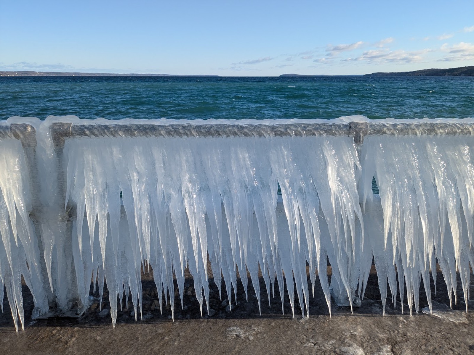 A shoreline railing encased in spray ice with a dense row of long icicles hanging from it, and open windblown bay water behind