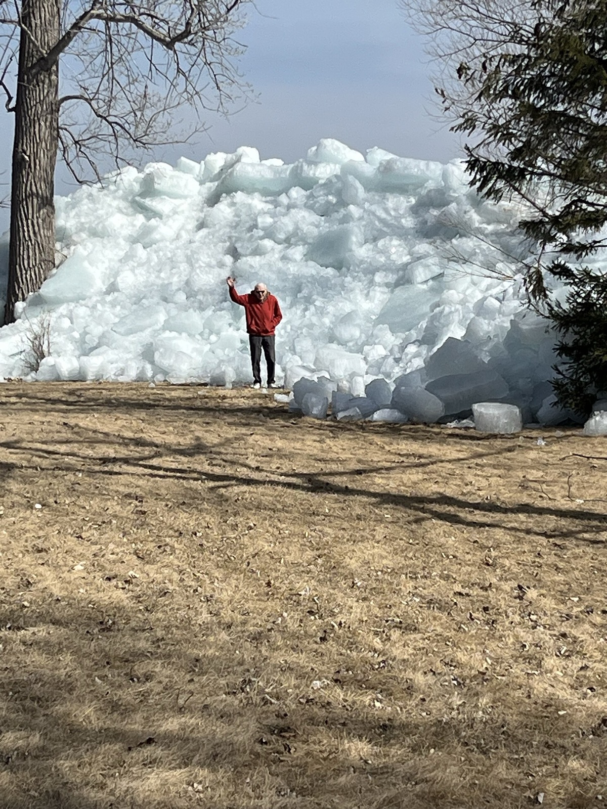 A person in a red jacket standing on brown grass at the foot of a wall of shattered lake ice piled far higher than they are