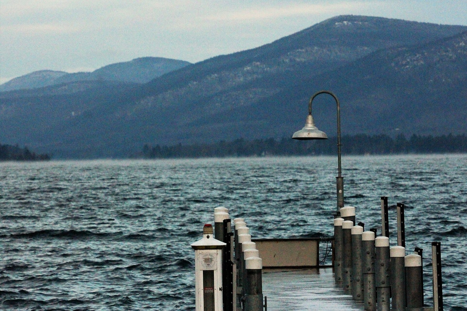 A wooden dock running out over still lake water in winter with snow on the boards and bare hills behind
