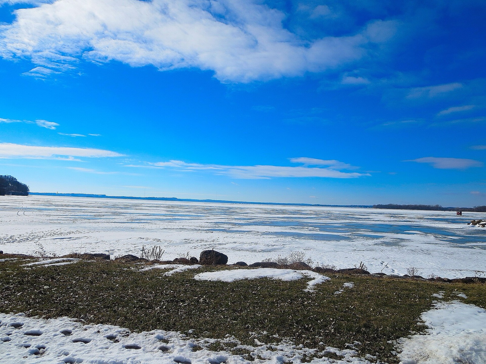 A frozen lake stretching to the horizon under a blue sky, with patchy snow and a rocky grass bank in the foreground
